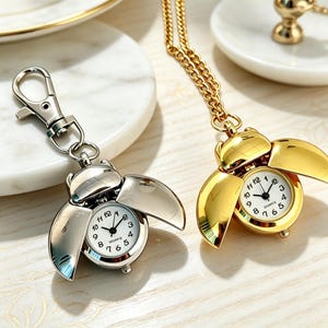 May include: Two beetle-shaped quartz watches with hinged wings that open to reveal a white clock face with black numerals. One watch is silver-toned with a lobster clasp, and the other is gold-toned and attached to a matching metal chain.