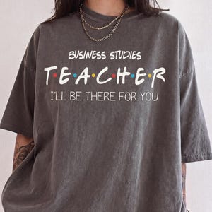 May include: A charcoal gray short-sleeve t-shirt featuring the white text BUSINESS STUDIES TEACHER with colorful dots between the letters of TEACHER and the white text I'LL BE THERE FOR YOU printed underneath.