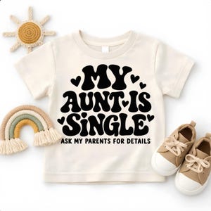 May include: A cream-colored short-sleeve t-shirt featuring bold black text that reads "MY AUNT IS SINGLE" with small heart accents and the additional text "ASK MY PARENTS FOR DETAILS" printed in a smaller, clean sans-serif font below the main graphic.