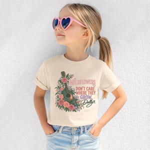 K&ouml;nnte beinhalten: Ein cremefarbenes Kurzarm-T-Shirt mit einer Grafik einer gr&uuml;nen Akustikgitarre, die von rosa und orangefarbenen Blumen umgeben ist. Der Text lautet "WILDFLOWERS DON'T CARE WHERE THEY GROW. Dolly" in einer Mischung aus rosa und schwarzer Schrift.