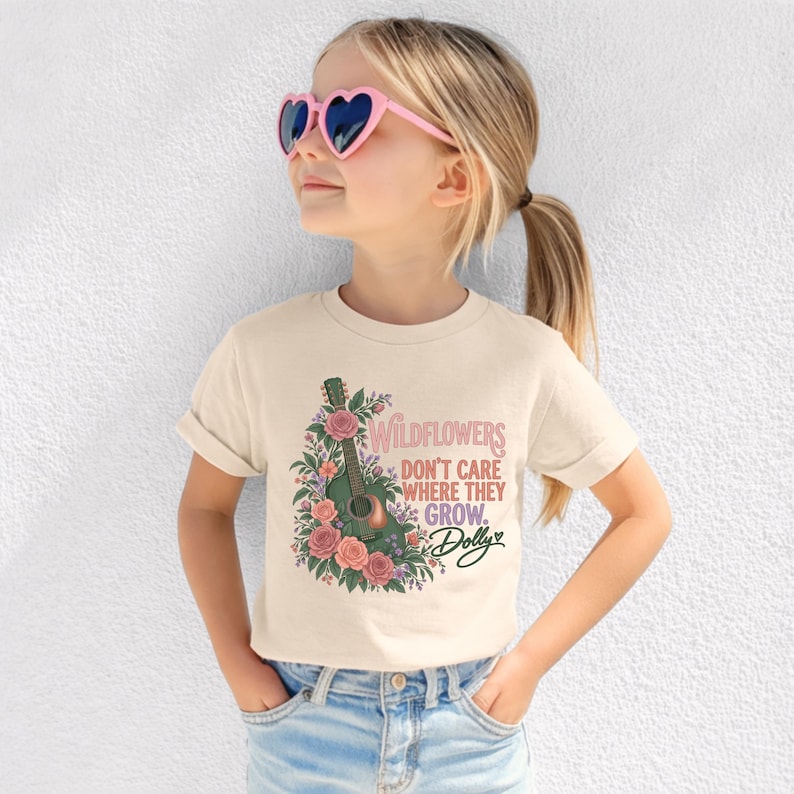 K&ouml;nnte beinhalten: Ein cremefarbenes Kurzarm-T-Shirt mit einer Grafik einer gr&uuml;nen Akustikgitarre, die von rosa und orangefarbenen Blumen umgeben ist. Der Text lautet "WILDFLOWERS DON'T CARE WHERE THEY GROW. Dolly" in einer Mischung aus rosa und schwarzer Schrift.