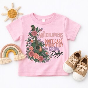 K&ouml;nnte beinhalten: Ein hellrosa T-Shirt mit kurzen &Auml;rmeln und einer Grafik einer gr&uuml;nen Akustikgitarre, umgeben von rosa und orangefarbenen Blumen. Der Text lautet: Wildflowers don't care where they grow. Dolly. Das T-Shirt ist mit Sonnen- und Regenbogendekor abgebildet.