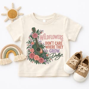 K&ouml;nnte beinhalten: Ein cremefarbenes Kurzarm-T-Shirt mit einer Grafik einer dunkelgr&uuml;nen Akustikgitarre, umgeben von rosa Rosen und violetten Wildblumen. Der Text lautet: Wildflowers don't care where they grow. Dolly, mit einem kleinen Herzsymbol.