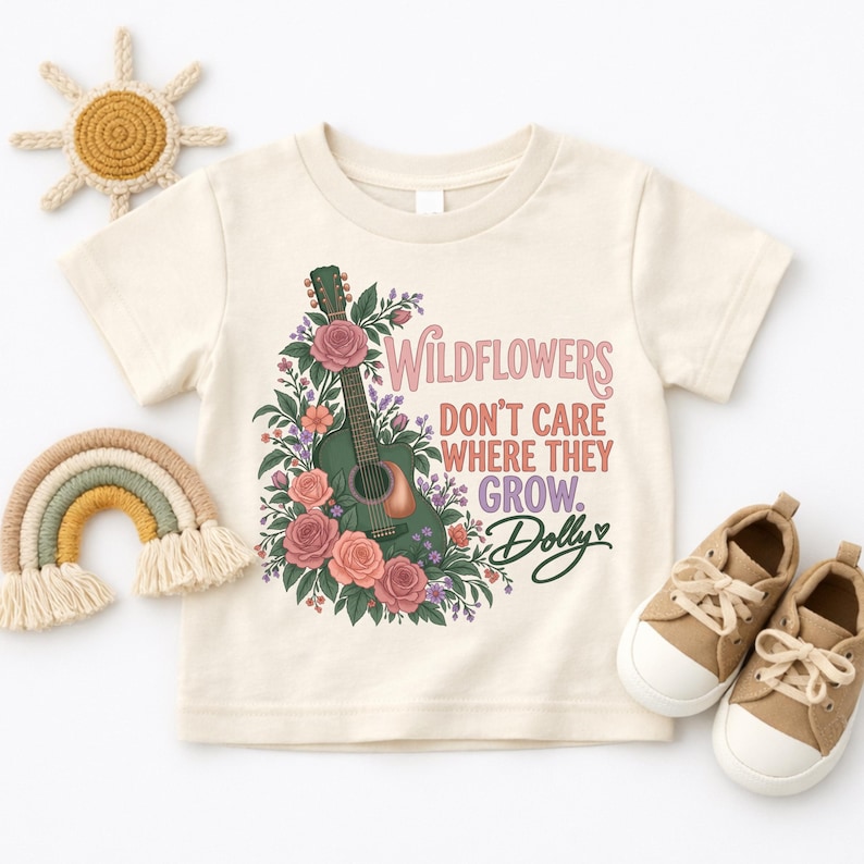 K&ouml;nnte beinhalten: Ein cremefarbenes Kurzarm-T-Shirt mit einer Grafik einer dunkelgr&uuml;nen Akustikgitarre, umgeben von rosa Rosen und violetten Wildblumen. Der Text lautet: Wildflowers don't care where they grow. Dolly, mit einem kleinen Herzsymbol.