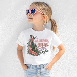 K&ouml;nnte beinhalten: Ein wei&szlig;es kurz&auml;rmeliges T-Shirt mit einer Grafik einer akustischen Gitarre, umgeben von rosa und roten Rosen sowie gr&uuml;nem Blattwerk. Der Text lautet: WILDFLOWERS DON'T CARE WHERE THEY GROW. Dolly, mit einem kleinen Herzsymbol daneben.