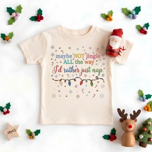 May include: A cream-colored short-sleeve t-shirt featuring the text maybe NOT jingle ALL the way I'd rather just nap in colorful letters, surrounded by small snowflakes, candy canes, and a string of multi-colored Christmas light bulbs.