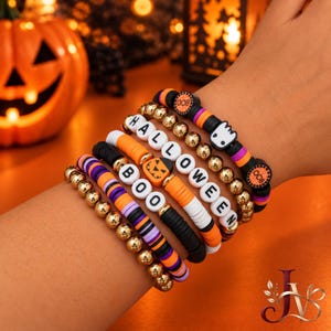 May include: A set of five stacked bracelets featuring black, orange, and purple clay beads, gold-toned metallic spheres, and letter beads spelling HALLOWEEN and BOO. The bracelets include a ghost charm and a jack-o'-lantern pumpkin bead.