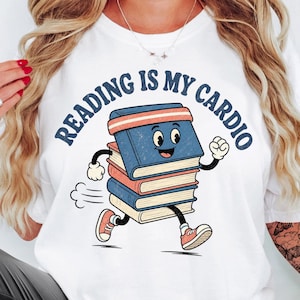 May include: A white t-shirt featuring a graphic of a stack of books with arms and legs running. The text above the books reads "READING IS MY CARDIO" in dark blue letters. The books are blue, red, and cream, and the character wears a headband.