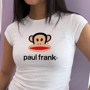 May include: A white short-sleeved t-shirt featuring the Paul Frank logo on the chest. The graphic displays a stylised monkey face with a tan complexion, black ears and hair, and a large red mouth, positioned above the black text paul frank.