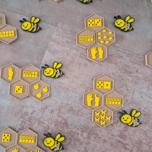 Op de afbeelding: Een set educatieve honingraatvormige tegels met gele iconen van handen, dobbelstenen, tientallenframes en diverse objecten, vergezeld door kleine geel-zwarte bijenfiguren. De tegels zijn in clusters gerangschikt en tonen numerieke weergaven.