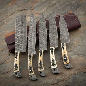 May include: A set of five knives with Damascus steel blades featuring a hammered texture. The handles are cream-colored with intricate black filigree patterns and brass rivets. The knives are arranged in a dark brown leather roll case.