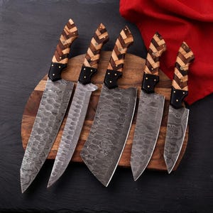 Könnte beinhalten: Ein Set aus fünf Küchenmessern mit gehämmerten Damaststahlklingen und Holzgriffen in einem hell- und dunkelbraunen Fischgrätenmuster. Die Messer haben schwarze Kropfe mit sichtbaren Nieten und liegen auf einem hölzernen Schneidebrett.