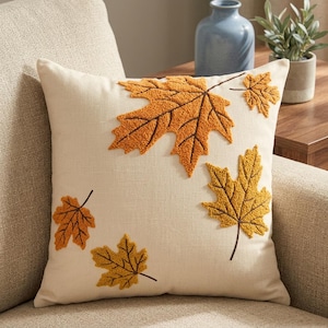 May include: A square throw pillow with a cream-colored fabric base featuring embroidered maple leaf designs in shades of orange and yellow. The leaves have a textured, raised appearance with visible stitching details, scattered across the front surface.