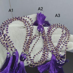 Peut inclure: Trois colliers de perles en pierre violette sur un fil fin doré, chacun terminé par un pompon en soie violet vif et des détails argentés. Les brins sont étiquetés A1, A2 et A3, avec de petites perles ovales violettes uniformes.