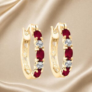 May include: A pair of gold hoop earrings featuring an alternating pattern of oval-cut deep red gemstones and small round clear stones set along the front curve of each hoop, secured with a hinged post closure mechanism.
