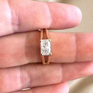 May include: A rose gold ring featuring a single rectangular radiant-cut clear gemstone set in a split-shank band. The center stone is held by four prongs, and the band design separates into two distinct metal lines that meet at the stone setting.