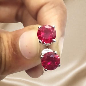 May include: A pair of stud earrings featuring round, faceted, deep red stones set in silver-toned metal prongs. The earrings have a classic circular design with a polished finish, showcasing the vibrant red color and reflective quality of the stones.