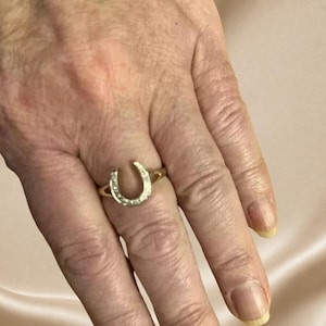 May include: A gold ring featuring a horseshoe-shaped centerpiece adorned with small, clear, sparkling stones. The horseshoe design is oriented with the opening facing downward, set on a thin, polished gold band that encircles the finger.