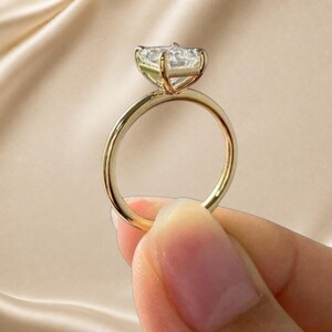 May include: A gold ring featuring a single, clear, rectangular-cut gemstone held in a four-prong setting. The band is thin, smooth, and polished, showcasing a metallic yellow finish that reflects light across its circular, continuous structure.
