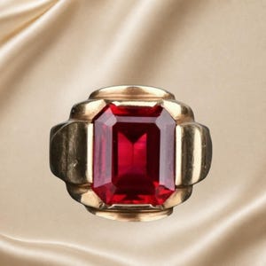May include: A gold ring featuring a large, rectangular, emerald-cut red gemstone set in the centre. The band has a wide, stepped, geometric design that frames the vibrant red stone, creating a bold and structured appearance on the finger.