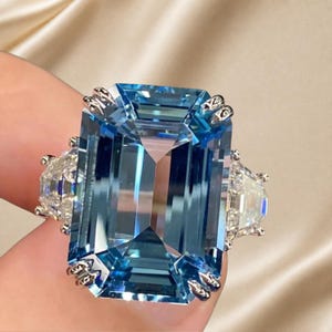 May include: A large emerald-cut blue gemstone ring set in a silver-toned metal band. The central blue stone is flanked by two smaller, clear, half-moon-shaped stones on either side, all secured with metal prongs in a three-stone ring design.