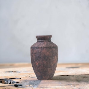 May include: A small, dark brown, mottled clay vase with a textured, weathered surface. The vase features a narrow neck with a subtle, rope-like embossed pattern encircling the shoulder, leading to a simple, flared rim at the top opening.