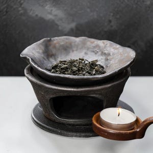May include: A dark, textured ceramic oil burner with a wide, shallow bowl containing dried green tea leaves. The burner features a central opening for a heat source and sits on a circular base. A small, lit white tea light candle rests nearby.