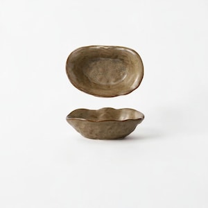 May include: Two small, oval-shaped ceramic bowls with an organic, wavy rim. The bowls feature a mottled, earthy brown glaze with a textured, uneven surface finish. The interior of the bowls shows a slightly lighter, speckled brown tone.