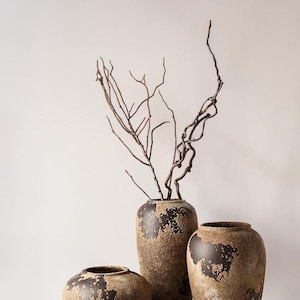 May include: Three ceramic vases with a textured, mottled brown and black finish. The vases have rounded bodies and wide openings. The tallest vase contains several thin, bare, dark brown branches extending upward from the opening.