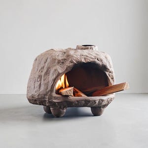 May include: A brown, rock-shaped outdoor fireplace with a textured, stone-like exterior. The structure features a wide, arched opening containing burning wood logs with visible orange flames and a small circular chimney vent on the top surface.