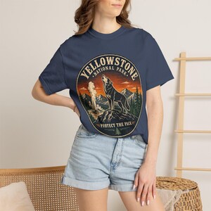 May include: A navy blue t-shirt featuring a circular graphic of a howling wolf on a rock, mountains, pine trees, and a geyser. The text reads YELLOWSTONE NATIONAL PARK and PROTECT THE PACK in a vintage-style font with an orange and blue sunset.