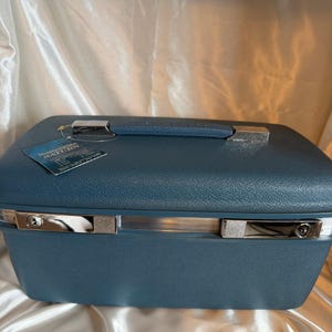 May include: A blue hardshell vanity case with a textured finish, featuring a top handle, silver-toned metal latches, and a key lock mechanism. A rectangular tag attached to the handle displays the text Samsonite SATURN in white lettering.