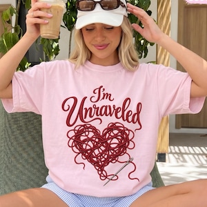 May include: A light pink short-sleeve t-shirt featuring the dark red text "I'm Unraveled" above a heart shape made of tangled yarn and a sewing needle. The shirt is worn with light blue and white striped shorts and mint green sneakers.