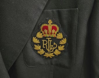 May include: A black blazer featuring a chest pocket with an embroidered crest. The crest displays a red and gold crown above the gold-stitched letters RL, all surrounded by a circular wreath of gold leaves on a dark fabric background.