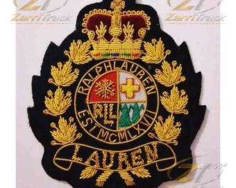 May include: A black embroidered patch featuring a gold crown at the top and a central circular crest divided into four quadrants with symbols. The text RALPH LAUREN EST MCMLXVII is stitched around the circle, with LAUREN on a banner below.