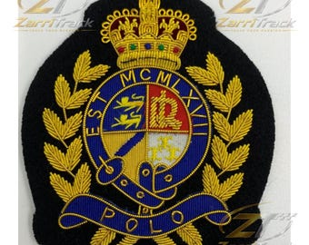 May include: A black embroidered patch featuring a gold crown at the top, surrounded by gold laurel leaves. A central blue circular border contains the text MCMLXVIII and POLO. The shield displays gold lions, a letter R, and fleur-de-lis symbols.