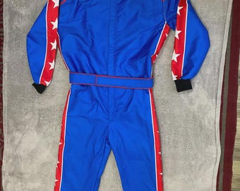May include: A blue racing suit featuring red side stripes on the arms and legs, decorated with white stars. The garment includes a high collar, a central zipper closure, a waist belt, and black cuffs at the wrists and ankles.