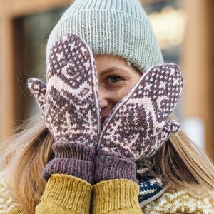 Könnte beinhalten: Ein Paar braun-cremefarbene Strickfäustlinge mit geometrischem Muster und Hirschmotiv. Die Fäustlinge haben ein geripptes Bündchen und werden zusammen mit einer hellgrünen Rippstrickmütze und einem senfgelben Pullover mit weißem geometrischem Muster getragen.