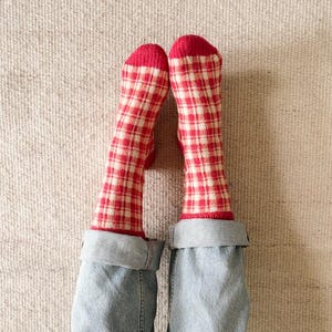 Könnte beinhalten: Ein Paar Socken mit rot-weißem Vichy-Karomuster und einfarbig roten Zehen- und Fersenbereichen. Die Socken werden zu einer hellblauen Jeans mit umgeschlagenen Säumen getragen, wobei der untere Teil der Beine und der gemusterte Stoff sichtbar sind.