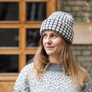 May include: A knitted beanie featuring a black and white grid pattern with a folded cuff. The beanie is worn with a grey and white sweater that has a repeating scalloped pattern across the chest and shoulders, constructed with a ribbed crew neckline.
