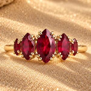 May include: A gold ring featuring five marquise-cut deep red rubies arranged in a row. The central ruby is the largest, flanked by two smaller rubies on each side, all secured in prong settings on a polished gold band that catches the light.