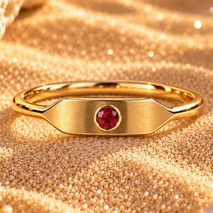 Könnte beinhalten: Ein goldener Ring mit einer flachen, rechteckigen Mittelplatte, in deren Mitte ein einzelner, kleiner, runder, leuchtend roter Edelstein eingefasst ist. Das Band ist glatt und poliert und zeigt ein schlichtes Design mit glänzender Oberfläche.