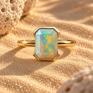 May include: A gold ring featuring a rectangular, emerald-cut opal gemstone with a vibrant play of color, including flashes of green, orange, and blue, set in a polished gold band with a smooth, minimalist metal setting surrounding the stone.