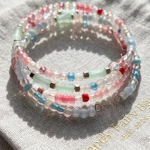 Peut inclure: Un bracelet multi-tours composé de perles translucides et opaques dans des tons rose, bleu clair, rouge et vert menthe, alternées avec de petites perles intercalaires métalliques cubiques et terminé par des boucles en fil métallique.