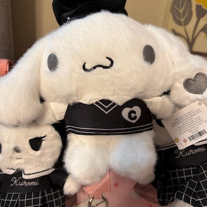 May include: A white plush toy with long ears, wearing a black hat and a black vest with a white heart and the letter C. Beside it are two smaller plush toys wearing black and white plaid skirts with the name Kuromi embroidered in white script.