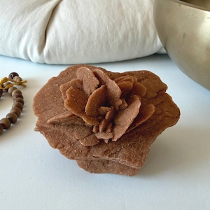May include: A desert rose crystal formation with a distinct, layered, flower-like structure. The mineral specimen features a sandy, earthy brown colour with a granular, textured surface composed of multiple thin, flat, and overlapping crystalline plates.