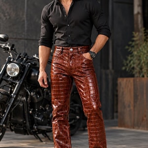 May include: A pair of glossy brown trousers featuring a textured crocodile skin pattern, paired with a black long-sleeved button-down shirt, black sunglasses, a black wristwatch, and black leather boots, standing near a black motorcycle.