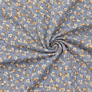 Puede incluir: Una tela de color azul claro con un patrón repetitivo de pequeñas flores de pétalos blancos y centros marrones. El material está fruncido en el centro, creando un efecto de espiral que resalta el estampado floral en la superficie textil.