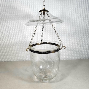 May include: A clear glass hanging lantern featuring a bell-shaped bowl with etched fern and floral patterns. The bowl is suspended by three metal chains attached to a matching glass canopy. A dark metal band with a star motif encircles the rim.