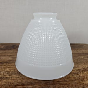 May include: A white glass lampshade with a textured, grid-like pattern. The shade has a flared, conical shape with a smooth, rounded base and a smaller, cylindrical top. The lampshade is designed to diffuse light.
